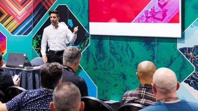Audience facing a presenter wearing dress clothes gesturing to a screen in front of a colorful mapping-themed backdrop