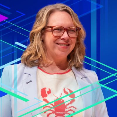 Smiling person wearing glasses, a light blue blazer, and a knit sweater with a lobster on it