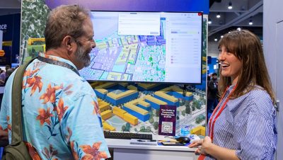 Two people standing at a conference kiosk viewing an interactive digital map on a large touchscreen display