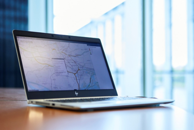 Opengeklapte laptop op een bureau met op het scherm een kaartweergave met datalagen en lijnen, gebruikt voor het analyseren van geografische informatie. Op de achtergrond zijn grote ramen zichtbaar met daglicht.