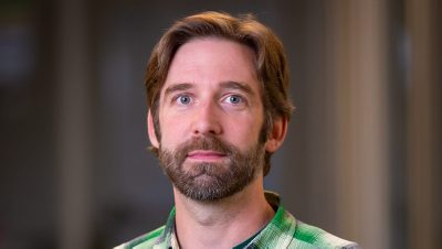 Portrait of Ryan Richardson in a green and blue plaid shirt with a blurred office background