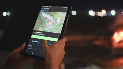 Hands hold a glowing tablet, its screen displaying a map with a fire symbol, against a backdrop of blurred lights