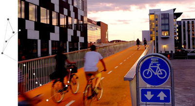 A bicycle bridge with 2 people riding bicycles and a bright blue traffic sign showing a bicycle and two arrows