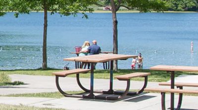 A couple sits at a wooden picnic table near a serene lake surrounded by lush greenery.
