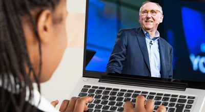 A woman is seated at a desk, using a laptop to view a virtual presentation. 