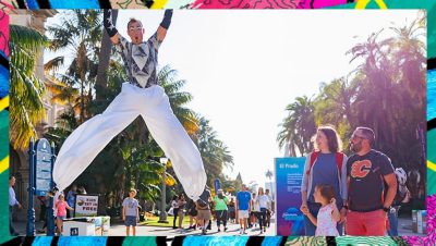 Performer in long white pants and a black and white top jumps into the air, hands up, while a family looks on