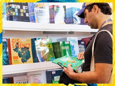 A person examining a book in front of shelves filled with colorful books and magazines including GIS Science titles