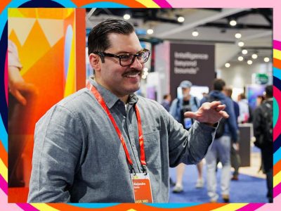 Esri expert with a mustache wearing a conference badge and glasses speaks at a booth with colorful branding