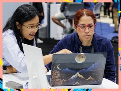 Two people collaborate at a table looking at laptops as one points at a screen with a mountain-and-moon design.