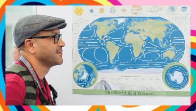 Smiling person wearing a cap and backpack stands beside a colorful educational world map poster showing global ocean currents, continents, and circular inset maps
