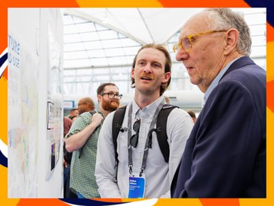 Jack Dangermond talking to an attendee in front of a map gallery exhibit in a bright exhibition hall