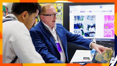 Two people at a conference booth review a map on a tablet while a large screen behind them displays GIS analysis visuals
