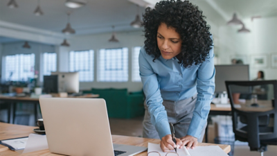 A businessperson standing at a desk writing on a notepad while looking at a laptop