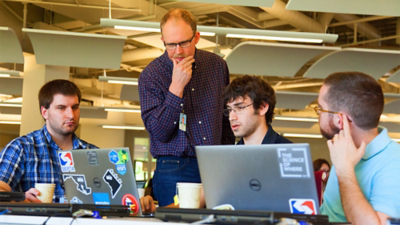 Three people sitting at a desk with laptops open and an instructor standing over them looking at their work