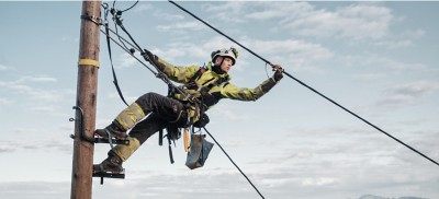 A lineworker, equipped with safety gear, diligently works on a utility pole by the ocean, with the sky and land in view.