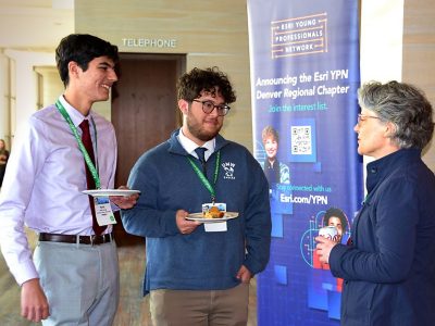 Two young people wearing conference lanyards and holding plates of food while talking to another person at an event 